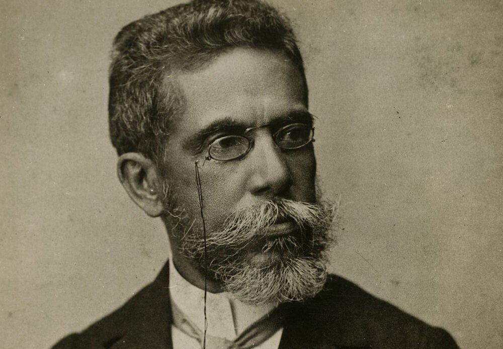 Retrato fotográfico em preto e branco de Machado de Assis, escritor brasileiro, com óculos redondos, barba grisalha e terno escuro