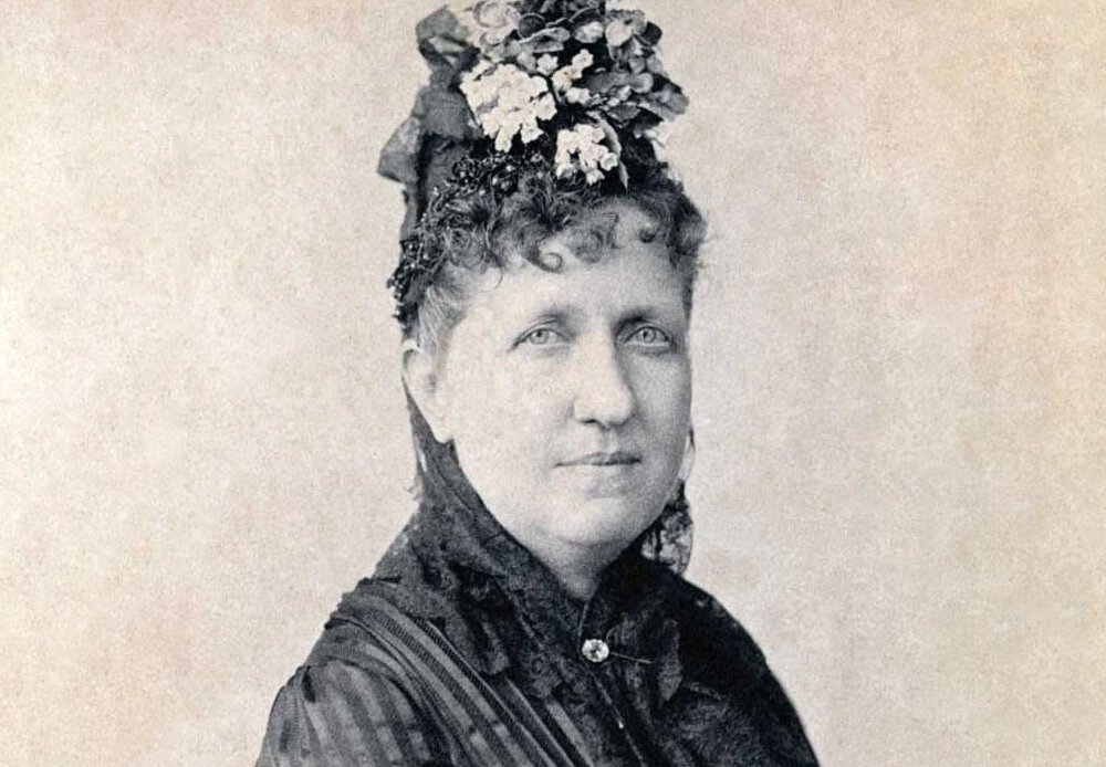  Retrato fotográfico em preto e branco da Princesa Isabel, vestida formalmente com chapéu adornado com flores e roupa escura de época