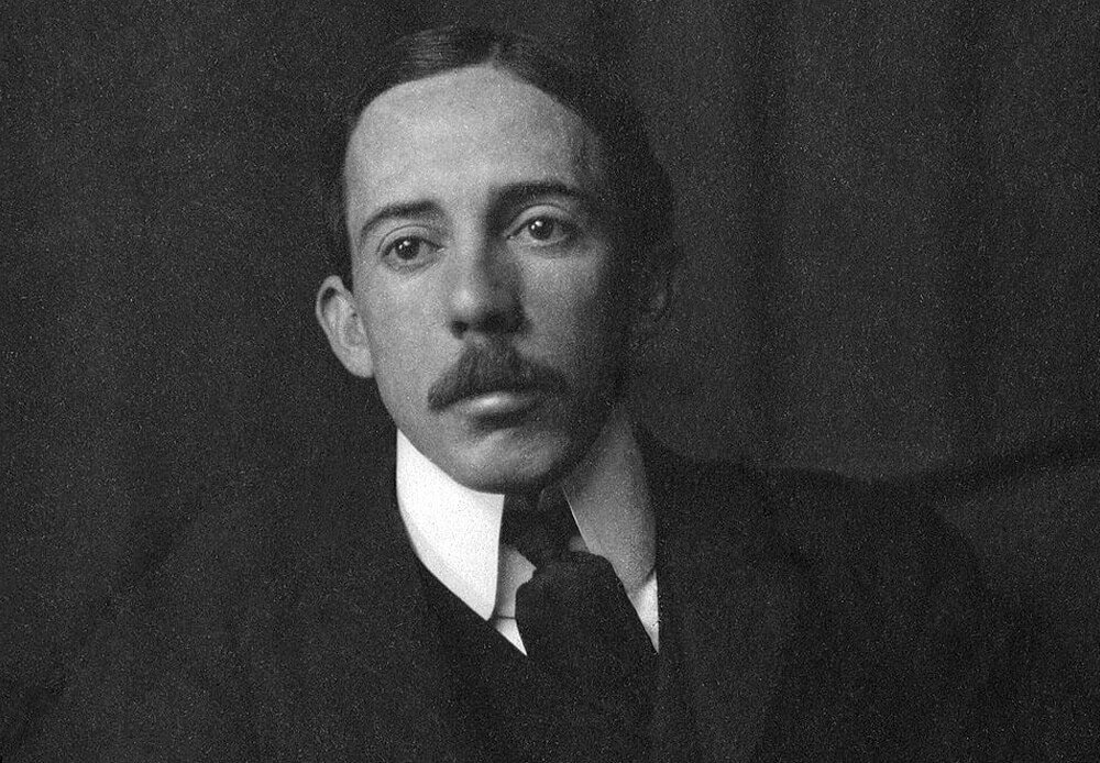 Retrato fotográfico em preto e branco de Alberto Santos Dumont, jovem, com bigode fino e terno escuro com gravata, olhar direto para a câmera
