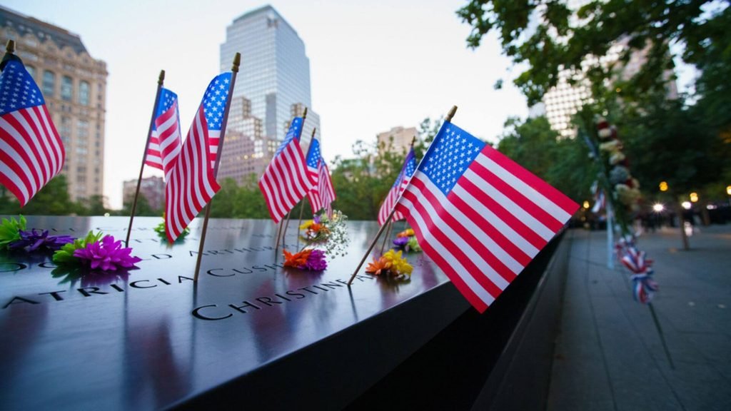 Nomes das vítimas gravados em bronze no Memorial 11 de Setembro em Nova York, com bandeiras dos EUA e flores deixadas em homenagem